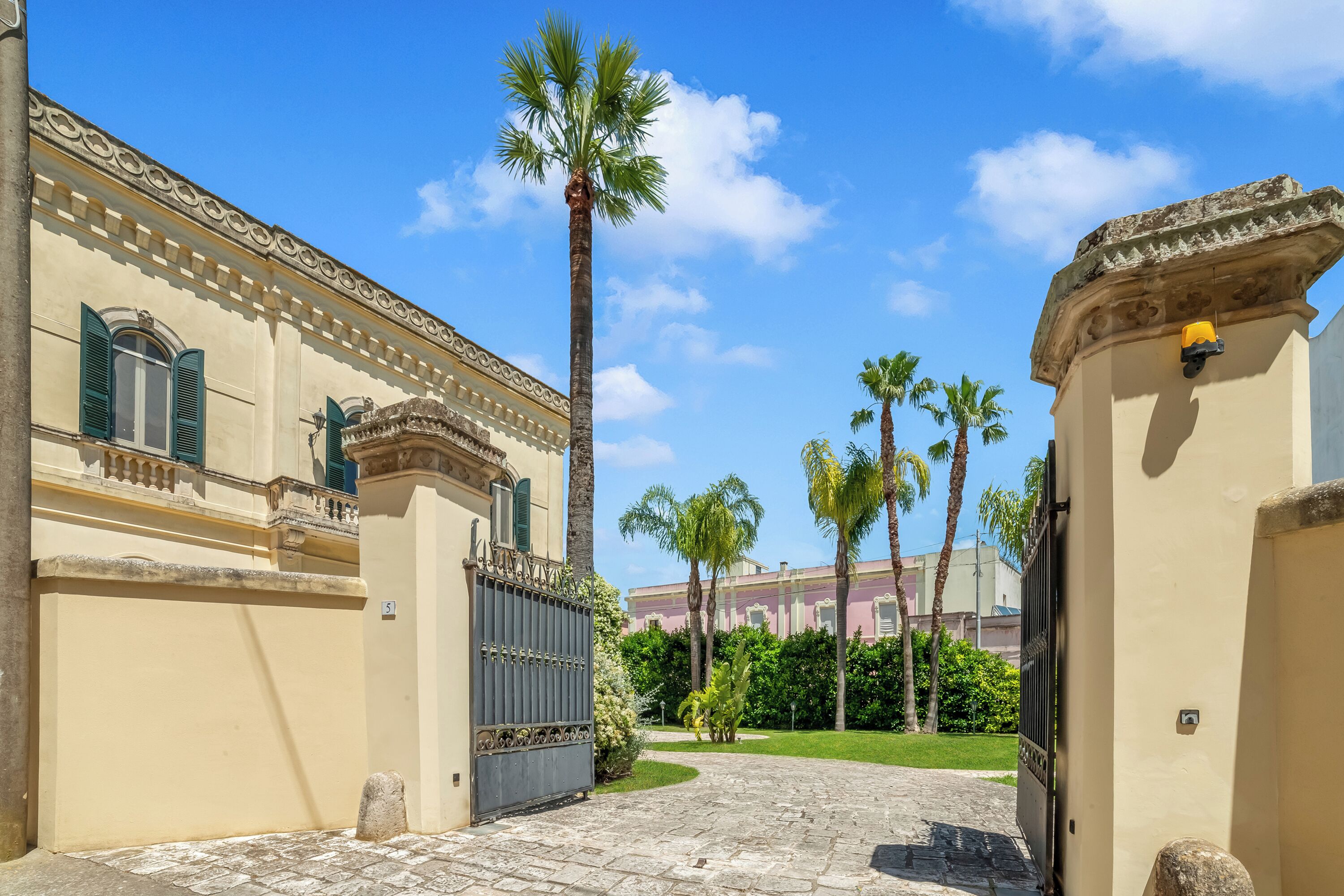 Une entrée fermée par un portail, un chemin pavé, des palmiers et un bâtiment à l’architecture classique.