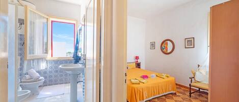 Una habitación de hotel con baño, una cama con cubrecama amarillo y una pequeña mesa con una silla.