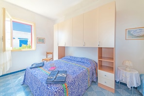 Ein Schlafzimmer mit einem Bett, einem Sessel, einem Kleiderschrank, einem kleinen Tisch mit Lampe und einem Fenster mit Blick auf ein Gebäude.