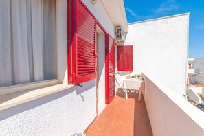 Ein schmaler Außenkorridor mit roten Fensterläden, einem weißen Tisch mit Tischdecke und einer kleinen Sitzecke.