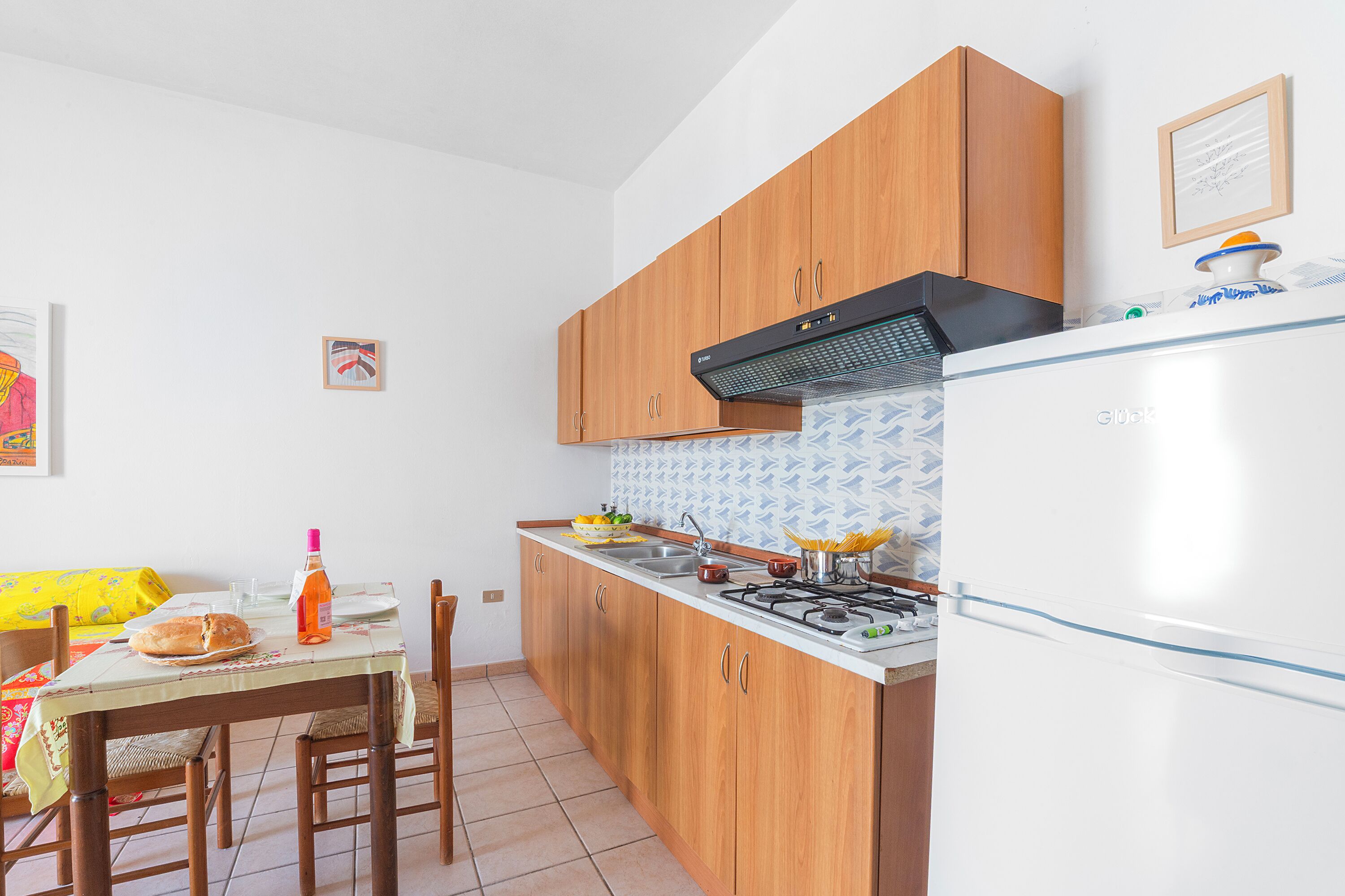 Eine Küche mit Holzschränken, einem Gasherd, einem weißen Kühlschrank und einem Esstisch mit Stühlen.