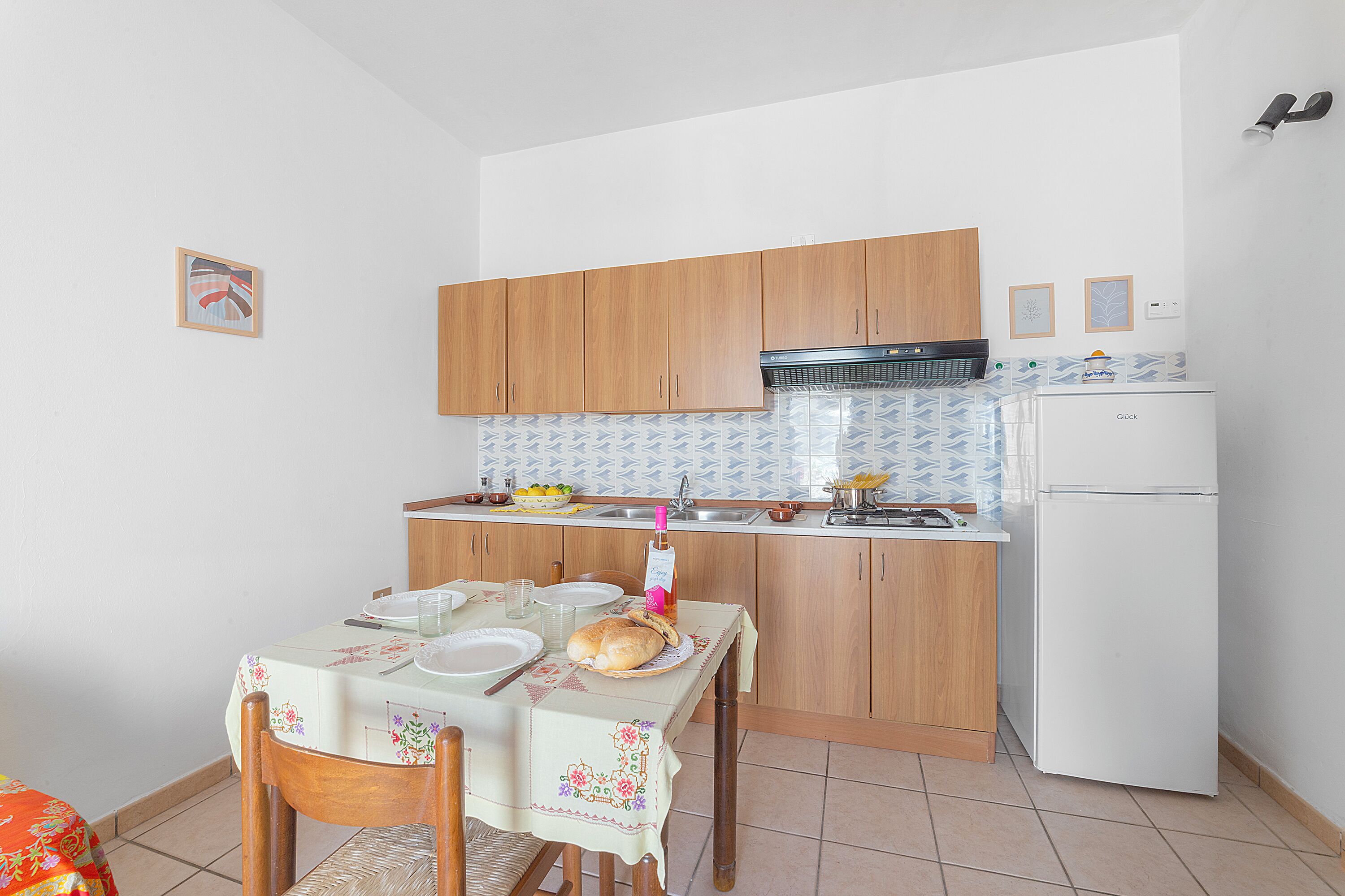 Een kleine keuken met houten kastjes, een witte koelkast en een eettafel gedekt voor twee personen.