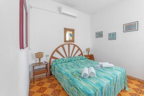 Ein Schlafzimmer mit einem hölzernen Bett, einem Nachttisch, einem Schreibtisch, einem Stuhl, einer Wand-Klimaanlage und einem gemusterten Boden.