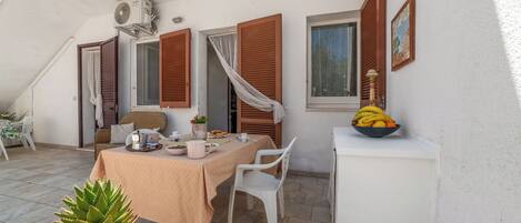 A dining area with a table set for two, a white chair, a small white cabinet with a bowl of fruit, and a window with brown shutters.