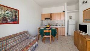 En kompakt opholdsstue med spisebord, stole, sofa, et tekøkken med skabe og et køleskab, samt et fjernsyn på et stativ.