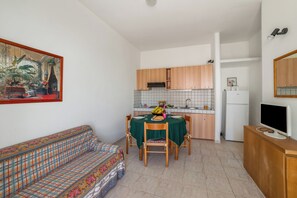 Ein kompakter Wohnbereich mit Esstisch, Stühlen, einer Couch, einer Küchenzeile mit Schränken und einem Kühlschrank, sowie einem Fernseher auf einem Schrank.