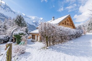 A wooden chalet with a sloped roof, surrounded by snow-covered trees and mountains in the background.