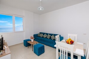 Una sala de estar moderna con un sofá azul, una pequeña mesa de madera con un florero, una silla blanca y una mesa de comedor con fruta.