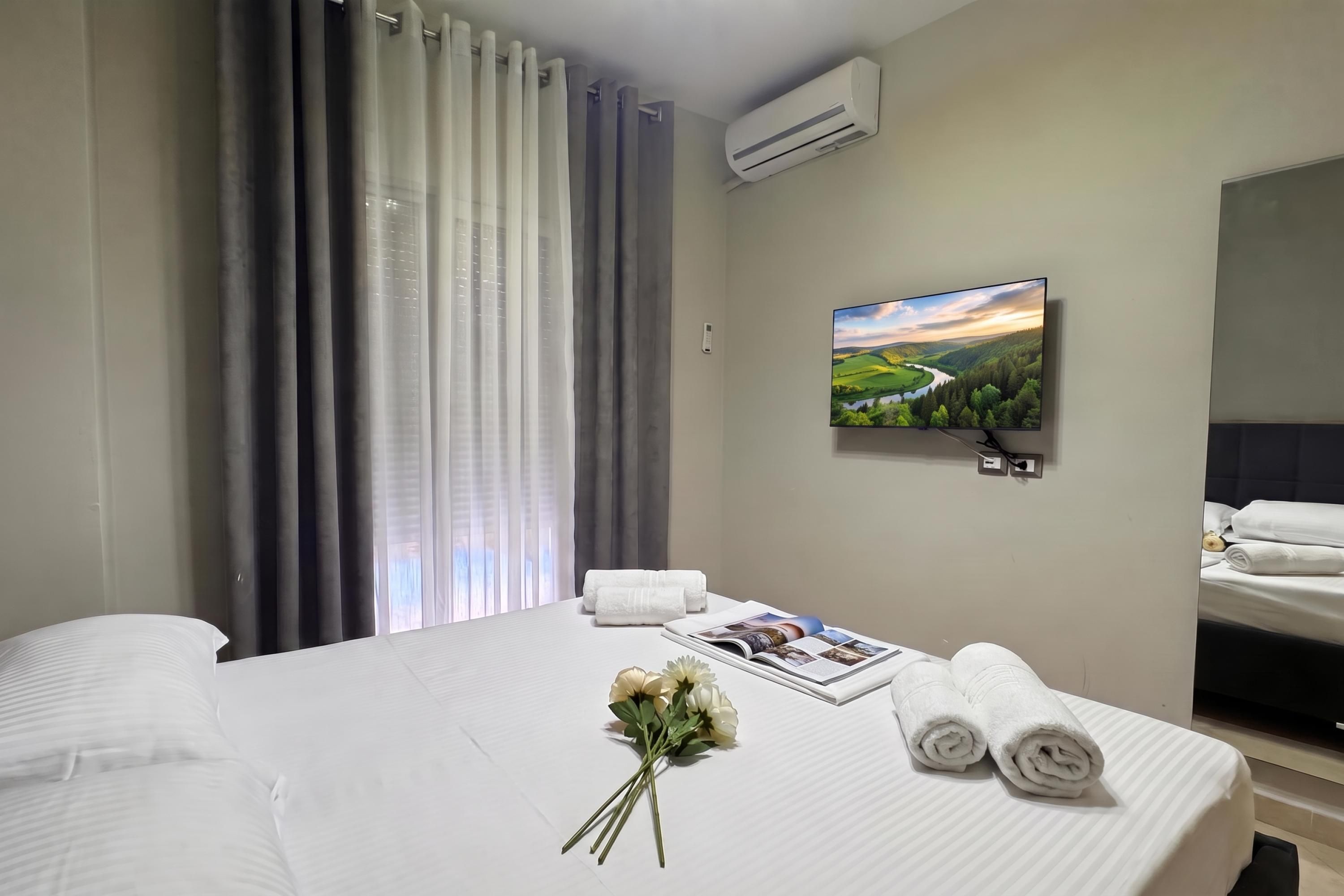 Ein Hotelzimmer mit einem Bett, einem Flachbildfernseher, einem Fenster mit Vorhängen und einem Blumenstrauß.