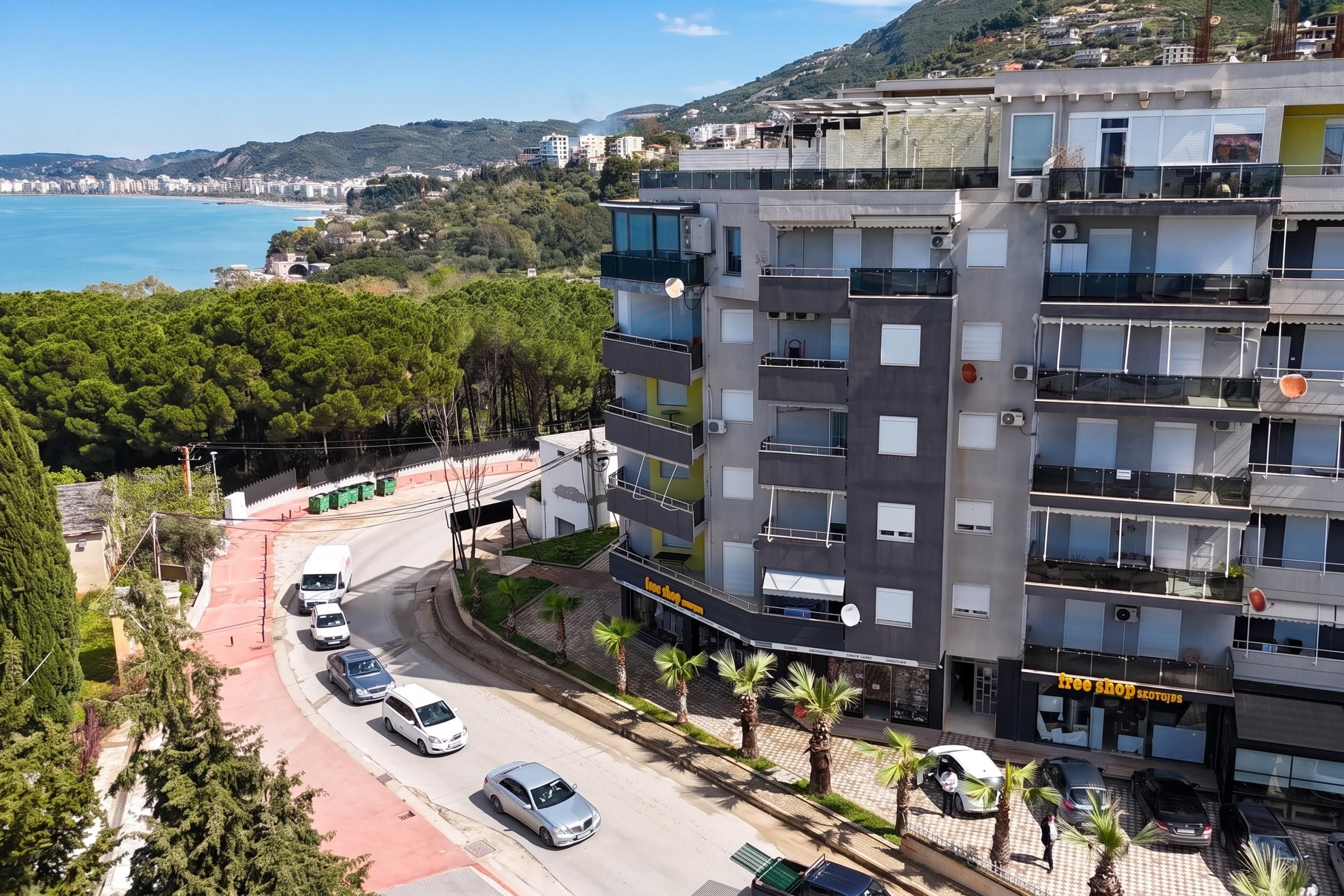 A multi-story building with balconies, a street with parked cars, and a view of the sea in the background.