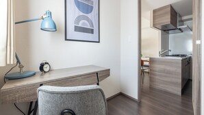 A modern room with a desk, chair, lamp, clock, and a kitchen area visible through an open door. - Dash Living Osaki (Shinagawa-ku)