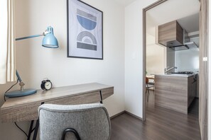 A modern room with a desk, chair, lamp, clock, and a kitchen area visible through an open door. - Dash Living Osaki (Shinagawa-ku)