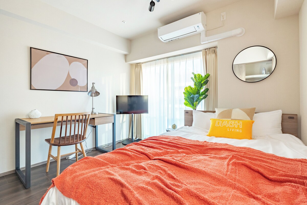 A hotel room with a bed, a desk with a chair, a television, a large window with curtains, a potted plant, and a wall-mounted mirror.