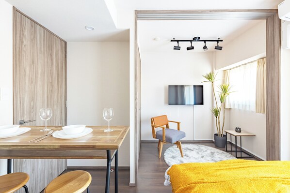 A modern room with a wooden dining table set for two, a single chair, a bed with a yellow blanket, a TV mounted on the wall, and a potted plant.