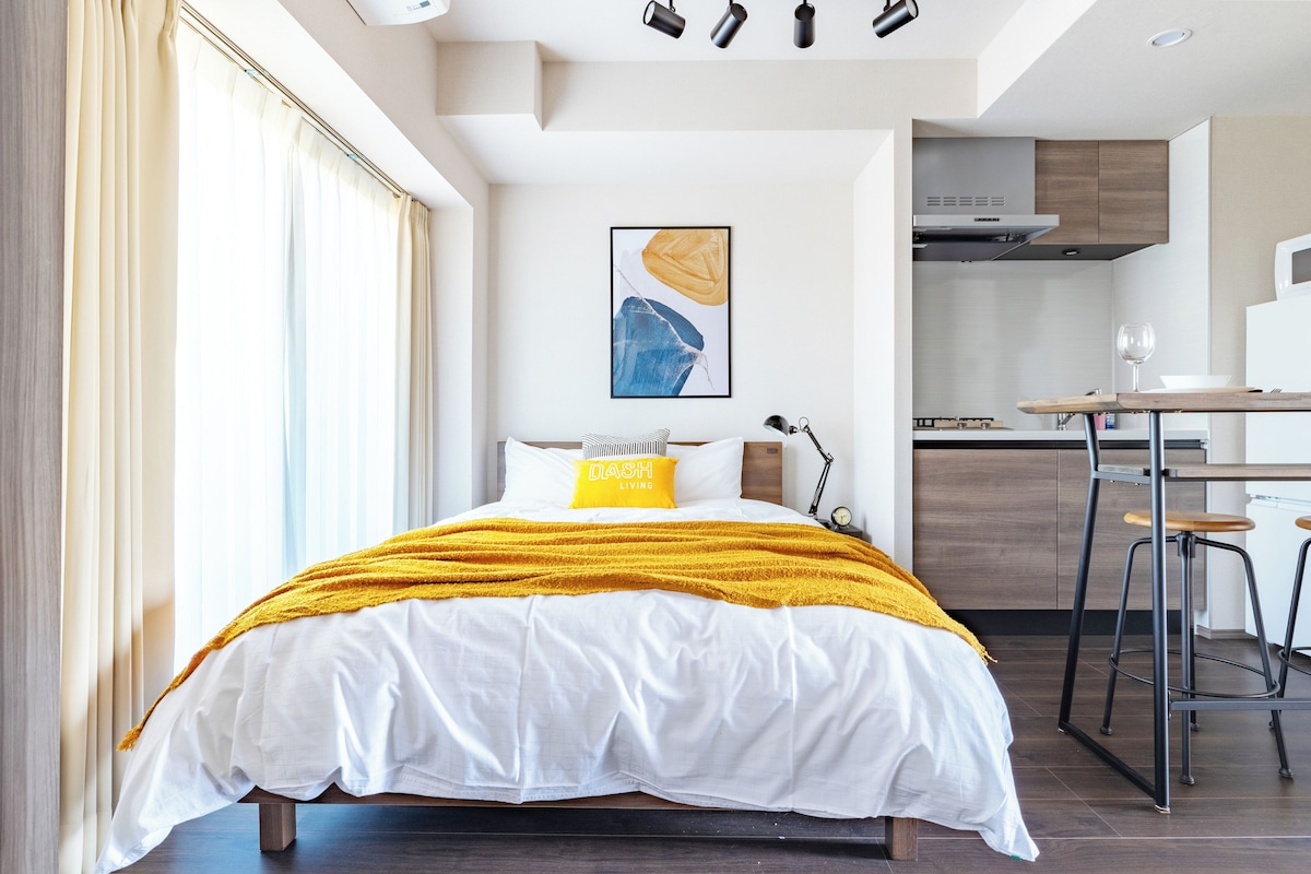 A modern hotel room with a bed, a desk, and a kitchen area.