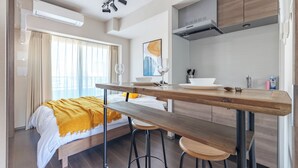 A modern hotel room with a bed, a wooden dining table, bar stools, a kitchen area, and a large window with curtains. - Dash Living Osaki (Shinagawa-ku)