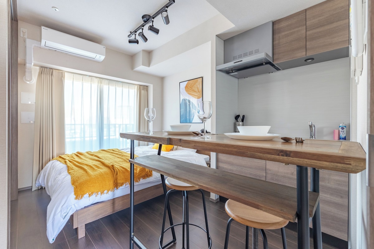 A modern hotel room with a bed, a wooden dining table, bar stools, a kitchen area, and a large window with curtains.