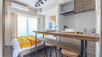 Una habitación de hotel moderna con una cama, una mesa de comedor de madera, taburetes de bar, una zona de cocina y un ventanal con cortinas.
