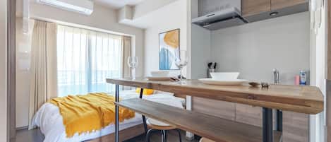 A modern hotel room with a bed, a wooden dining table, bar stools, a kitchen area, and a large window with curtains.