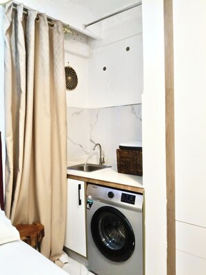 A compact kitchen with a washing machine, a sink, and a small wooden stool. - Prime Kilimani Location,5 mins Walk to Yaya Centre, Mersen Studio (Nairobi)