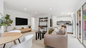 A modern living room with a sofa, a dining table, and chairs. There is a kitchen area with cabinets and appliances.