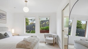 A bedroom with a bed, a chair, a bedside table, a mirror, and windows with a view of greenery. - Stylish West Hobart Villa (West Hobart)
