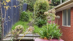 A garden with a variety of plants, a concrete pathway, and a brick building with a window. - Stylish West Hobart Villa (West Hobart)