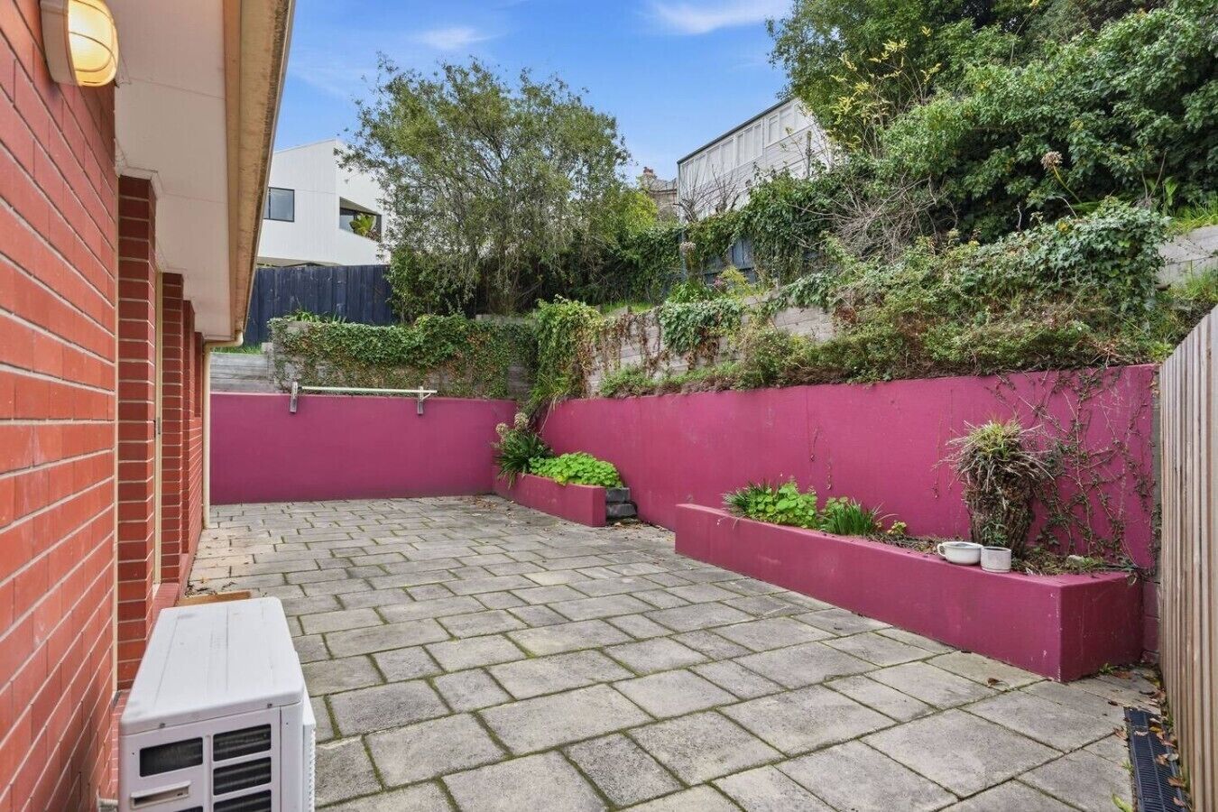 A paved patio area with a red wall and planters, a white air conditioning unit, and a brick wall.