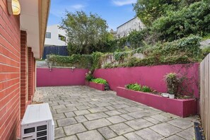 A paved patio area with a red wall and planters, a white air conditioning unit, and a brick wall. - Stylish West Hobart Villa (West Hobart)