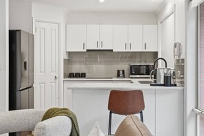 Una cocina compacta con armarios blancos, un refrigerador de acero inoxidable, microondas y cafetera.