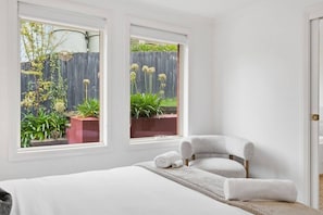 Ein Schlafzimmer mit einem Bett, einem Sessel und zwei Fenstern mit Blick auf einen Garten.