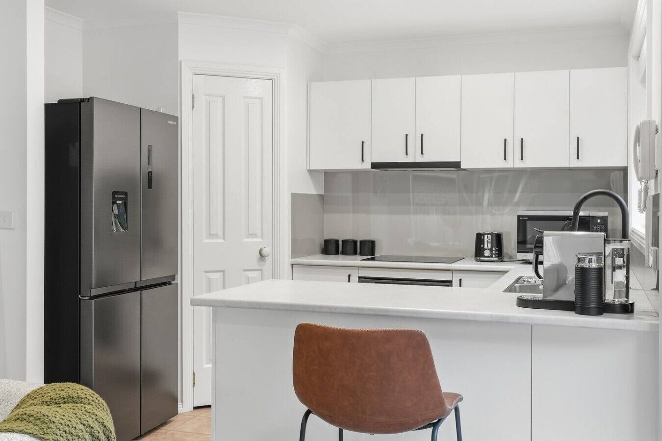 A compact kitchen with white cabinets, a stainless steel refrigerator, a coffee machine, and a single chair.