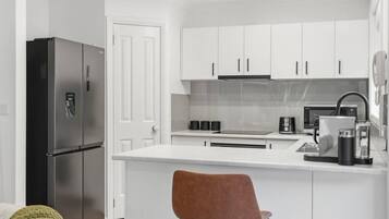 A compact kitchen with white cabinets, a stainless steel refrigerator, a coffee machine, and a single chair.