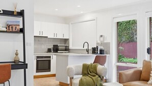 A modern kitchen with white cabinets, a microwave, and a coffee maker. There is a dining area with a table and chairs, and a seating area with a sofa and a chair. - Stylish West Hobart Villa (West Hobart)