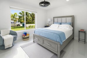 Ein modernes Schlafzimmer mit Bett, Sessel, Nachttisch und Fenster mit Blick auf Palmen und parkende Autos.