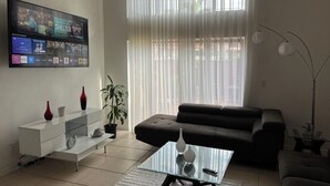 A living room with a flat-screen TV, a sofa, a glass coffee table, and a potted plant.