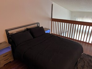 Een bed met hoofdeinde, een nachtkastje met lamp, een houten vloer en een raam met luxaflex.