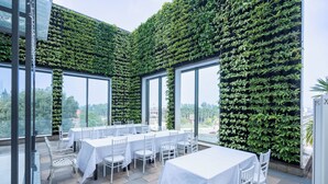Um restaurante com mesas e cadeiras brancas, grandes janelas e uma parede coberta de folhagem verde.