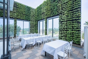 A restaurant with white tables and chairs, large windows, and a wall covered in green foliage. - Prime Hotel Thai Nguyen (Phan Đình Phùng)