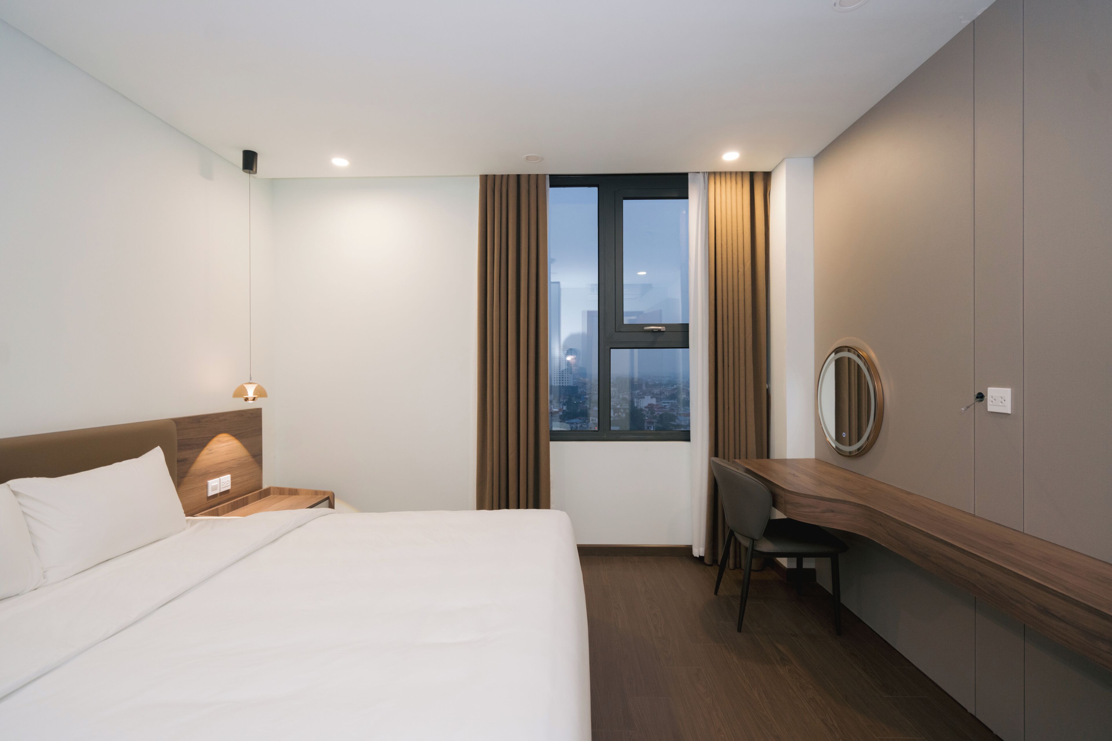 A hotel room with a bed, a desk, a chair, a window with curtains, and a view of the cityscape.