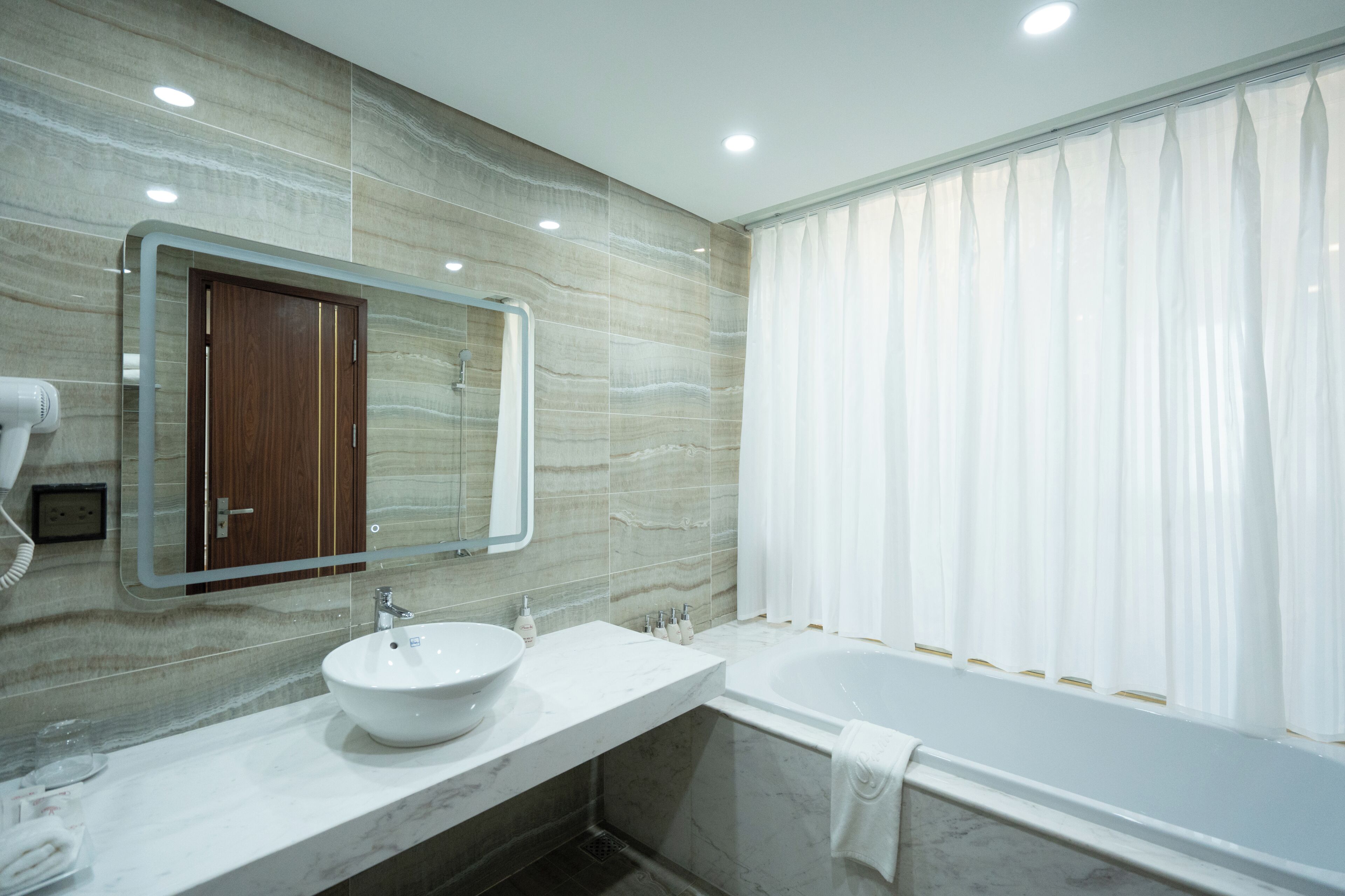 A modern bathroom with a marble vanity, a large mirror, a bathtub, and a window with curtains.