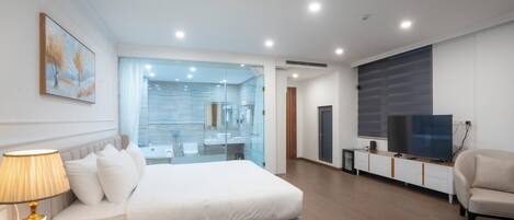 Quarto de hotel moderno com uma cama grande, mesa de cabeceira, luminária, TV e banheiro com paredes de vidro.
