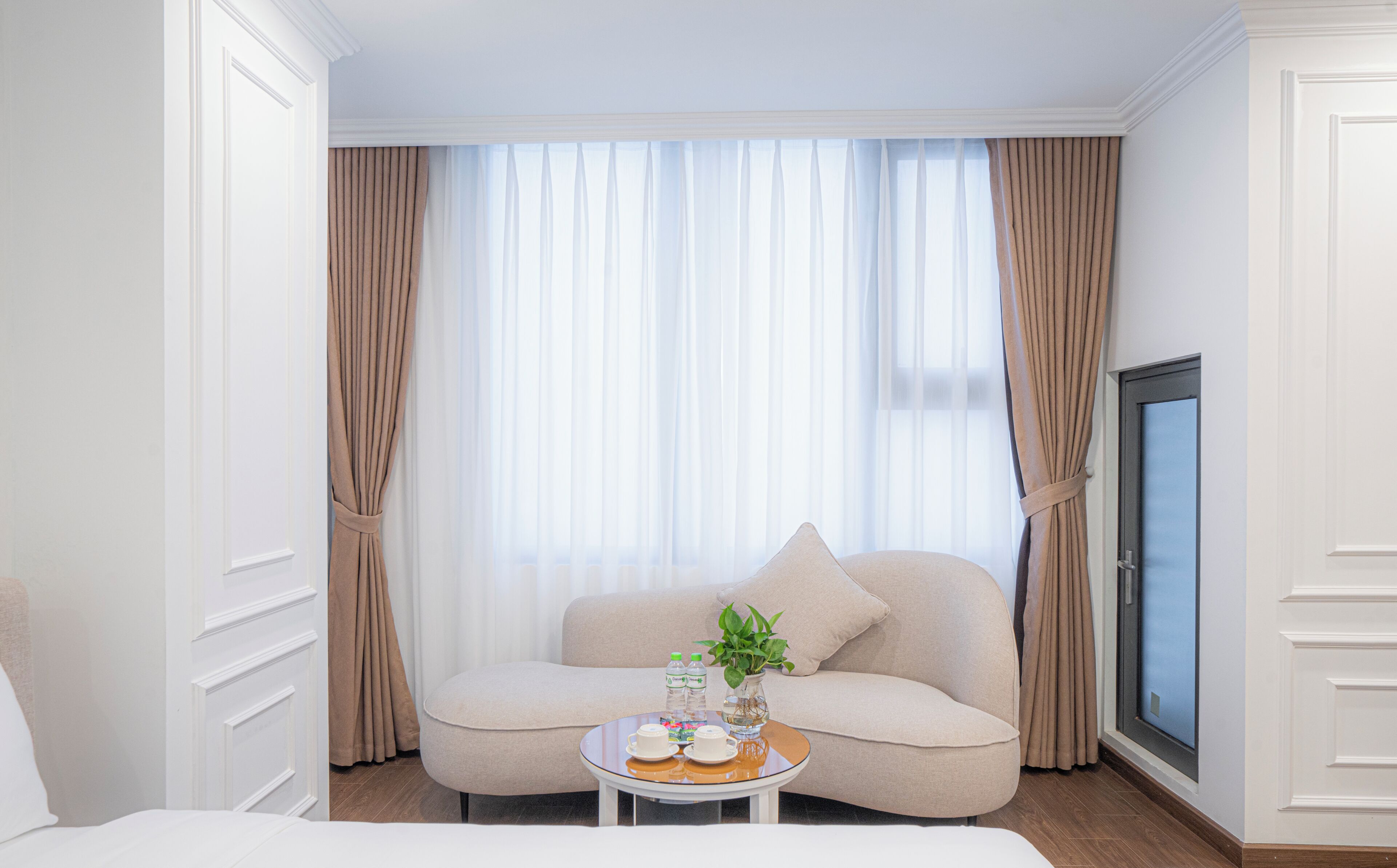 Habitación de hotel con sofá, una pequeña mesa redonda con un florero y dos botellas, y un ventanal con cortinas.