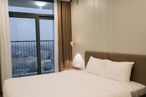 Habitación de hotel con una cama grande, una mesita de noche, una lámpara, una ventana con cortinas y vistas a la ciudad.