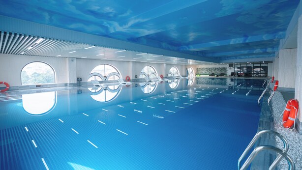 Piscina interna com água cristalina, teto com design em formato de nuvens e janelas ao longo das paredes.