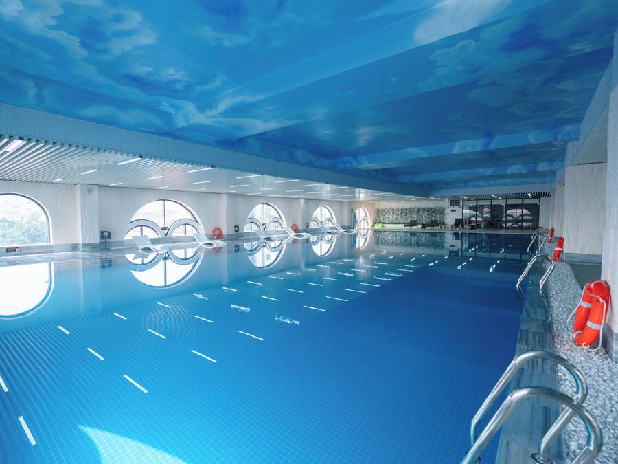 An indoor swimming pool with clear water, a ceiling with a cloud design, and windows along the walls.