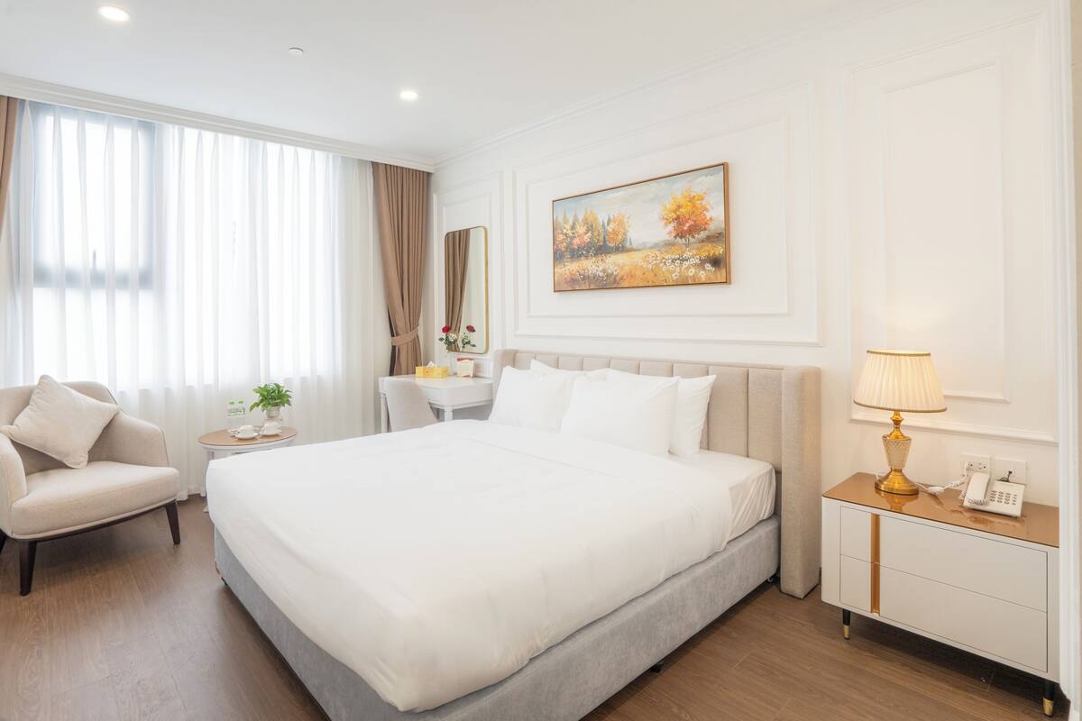 A hotel room with a large bed, a chair, a bedside table, a lamp, and a painting on the wall.