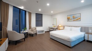 Quarto de hotel com cama, escrivaninha, cadeira e uma janela grande.