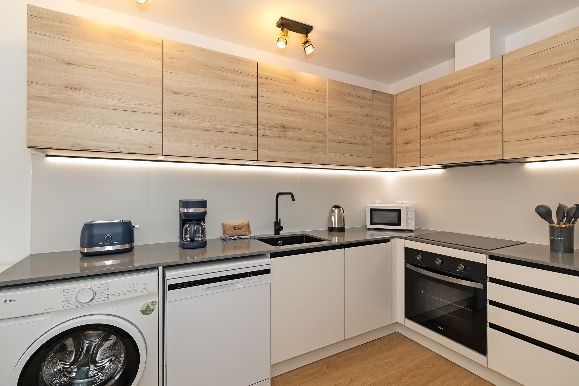 A modern kitchen with wooden cabinets, a dishwasher, a coffee maker, a kettle, a microwave, and a built-in oven.