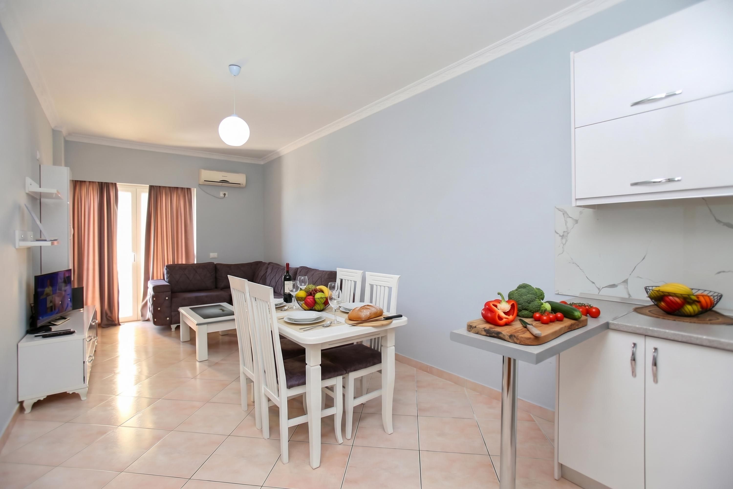 A modern living room with a sofa, a dining table set for two, and a kitchen area with a fruit bowl and vegetables.